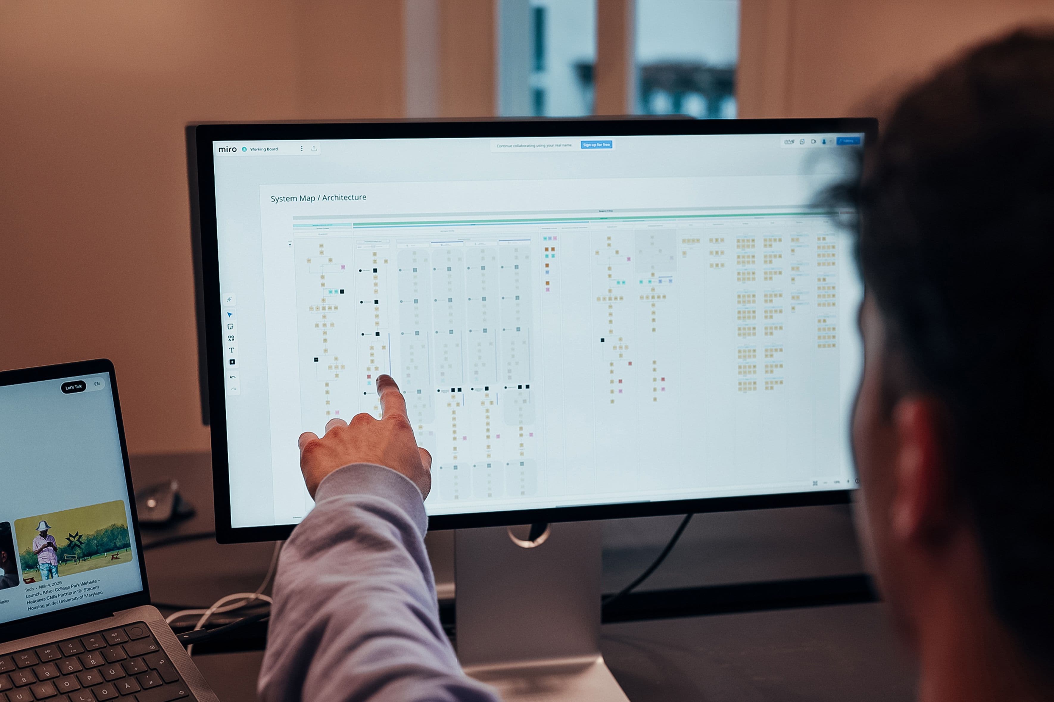Ein Mann zeigt mit dem Finger auf einen großen Computermonitor, auf dem eine komplexe Systemarchitektur eines Produktes in der Software Miro dargestellt ist.