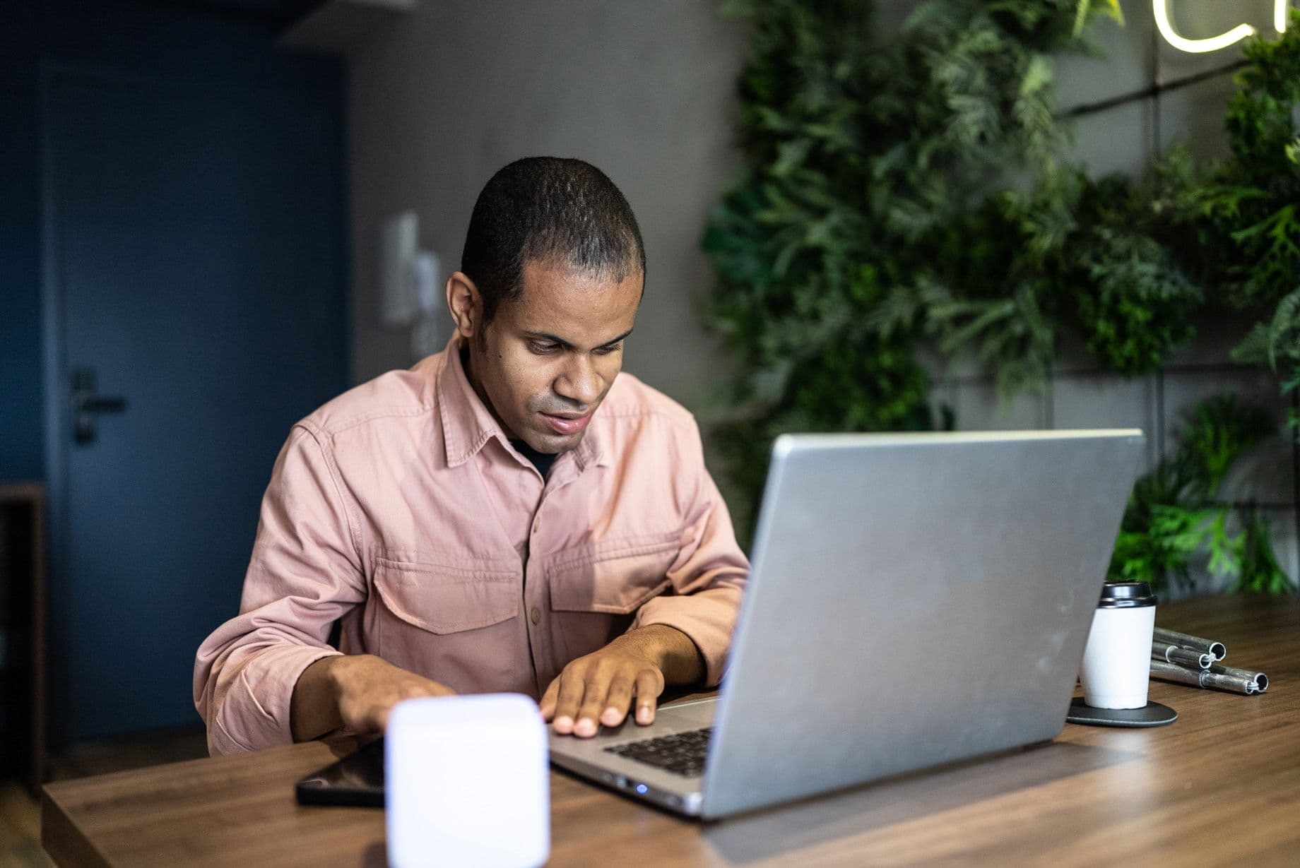 A visually impaired man using a laptop with assistive technology, demonstrating accessibility in digital workspaces. A coffee cup and office items are on the desk, with greenery in the background.
